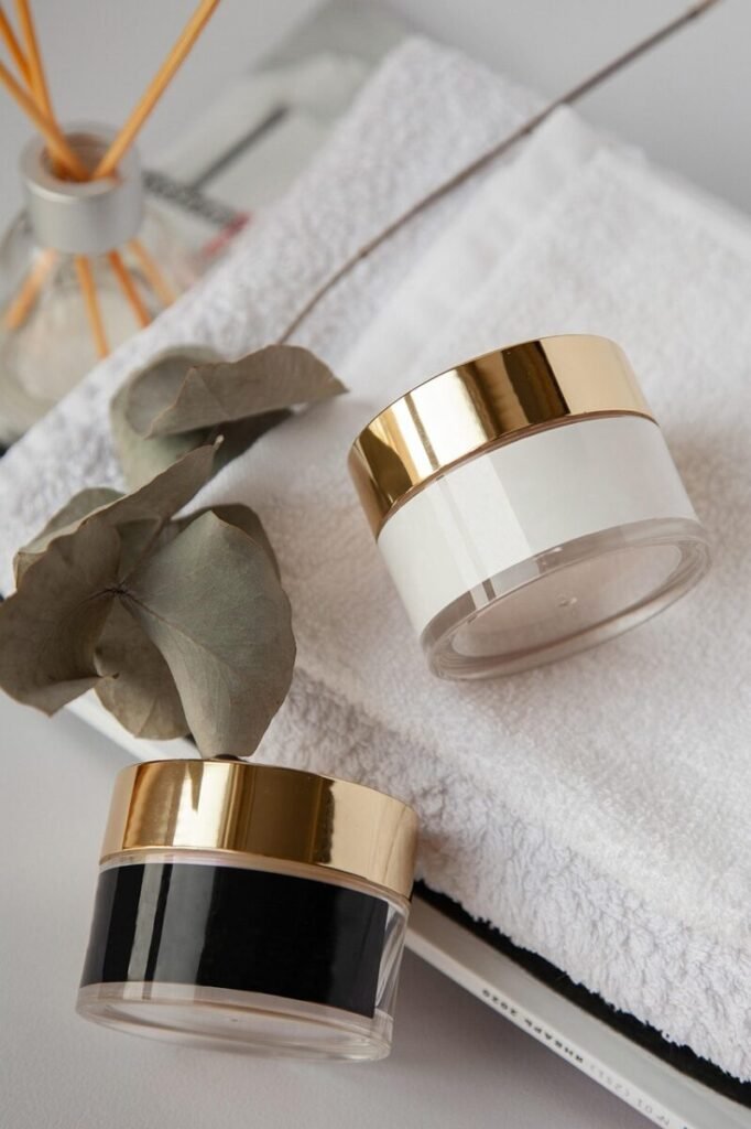 Two cream jars with gold lids resting on a folded white towel beside dried eucalyptus